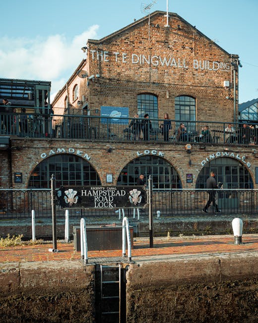A multi-storey brick building with a large sign reading 'The Tedingwall Building' at the top, situated near a canal in Hampstead. The ground floor features large arched windows and signage for the 'Hampstead Road Lock,' with a black metal railing running along the edge of a walkway. Several people are visible on the upper level, some walking and others standing by the railing, possibly observing the surroundings or preparing for a home relocation. The foreground shows a paved area with bollards and a small set of stairs leading down to the canal. The scene is captured in daylight with a partly cloudy sky, and the environment suggests an active area involved in moving or transportation activities, aligning with the theme of house removals and furniture transport facilitated by Hampstead Movers.