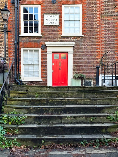The image shows the front entrance of a red-brick residential building identified as Holly Mount House, with a white sign above the window displaying its name. The building has three visible white-framed sash windows on the upper floors, two closed on the left and one open on the right. The ground floor features a bright red main door with a small window at the top, set within a white frame. Leading up to the door are concrete steps that are weathered and moss-covered, with some greenery growing between the cracks. On either side of the steps, black metal railings section off the entrance area; the left side has a vintage-style lamp post, and the right side has part of an iron fence. To the right of the door, there is a small green potted plant on a white stone surface, and a folding metal chair is partially visible behind the fence. The scene suggests the house is part of a typical London street, and the overall context hints at a location where house removals and furniture transport might be carried out, with the exterior setting captured during daytime with natural lighting.