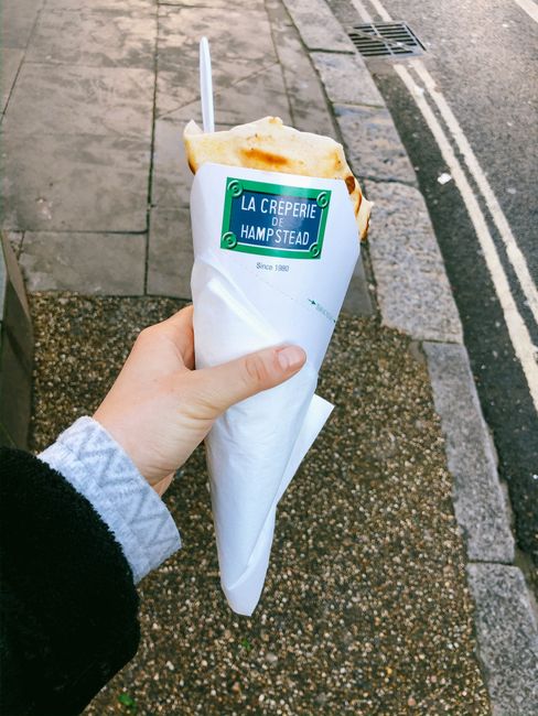 Close-up image of a person holding a wrapped crepe from La Creperie de Hampstead, with a white paper cone and a wooden stir stick inside. The person's hand is visible, dressed in a grey and black sleeve, and they are standing on a sidewalk featuring paved stones and a street with a metal drain cover and curb visible in the background. The crepe appears freshly cooked, with a golden-brown surface, and is partially inside the paper wrapping, which is branded with the creperie’s green and blue logo, referencing Hampstead. The scene suggests a casual moment during a walking or shopping trip on Hampstead High Street, with the visible environment indicating a typical urban setting suitable for house removals or relocation planning discussions, as highlighted in Hampstead Movers’ services related to home moving, packing, and transportation logistics.