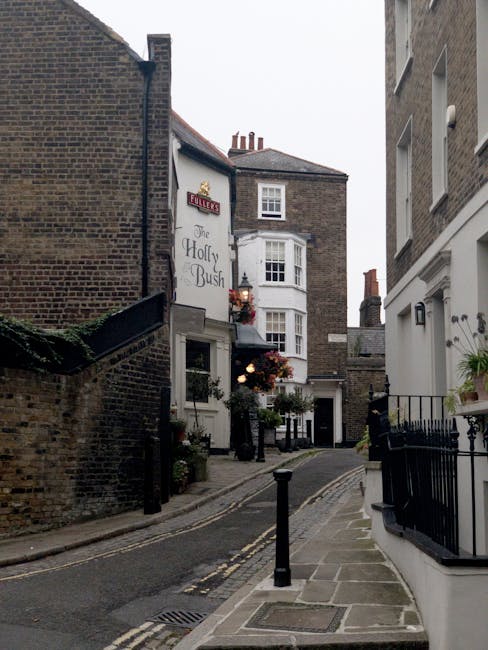 A narrow street in Hampstead lined with traditional red-brick and stone buildings, featuring a mix of shops, cafes, and pubs. On the left side, a dark blue pub facade with large glass windows and hang