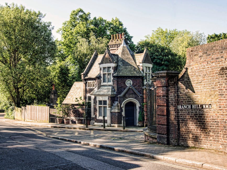 A residential street scene showing a historic, Victorian-style house with intricate brickwork, gabled roofs, multiple chimneys, and bay windows, situated behind a brick gate with a metal fence. The house is surrounded by tall, leafy trees and a wooden fence along the pavement. The scene is well-lit with natural daylight, illustrating a quiet neighbourhood in NW3 near South End Green, where Hampstead Movers may assist with home relocations. The road in front has a gentle curve with a double yellow line, and a street sign reads 'Branch Hill N.W.3' attached to the brick wall on the right, indicating the location. The image captures the environment typical of house removals or furniture transport logistics involving the careful handling of items through the gate and driveway, highlighting the importance of professional packing and moving services.