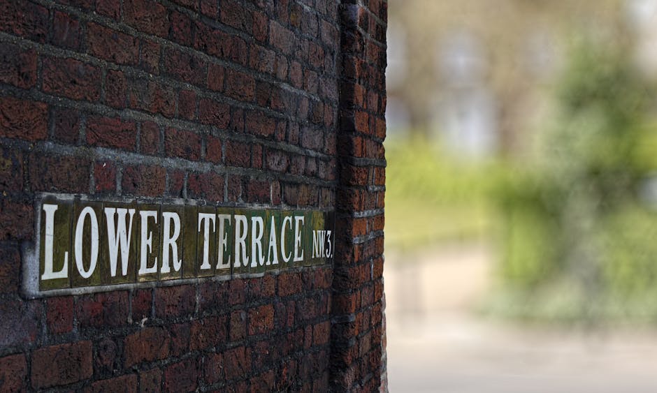 A close-up view of a brick wall featuring a rectangular street sign reading 'LOWER TERRACE NW3' in white uppercase letters on a green background. The sign is mounted on a dark red brick surface with visible mortar joints. In the background, a slightly out-of-focus view of a pedestrian sidewalk and green foliage can be seen, suggesting an outdoor setting near a residential or mixed-use area in Hampstead. The scene is well-lit with natural daylight, and the wall appears to be part of a building facade adjacent to a walkway used for home relocation or moving services. This image is relevant to house removals, transport, and packing operations, highlighting the typical environment encountered during local house moves near South End Green. Hampstead Movers, a professional removal company, would handle such environments during furniture transport or packing and moving processes.