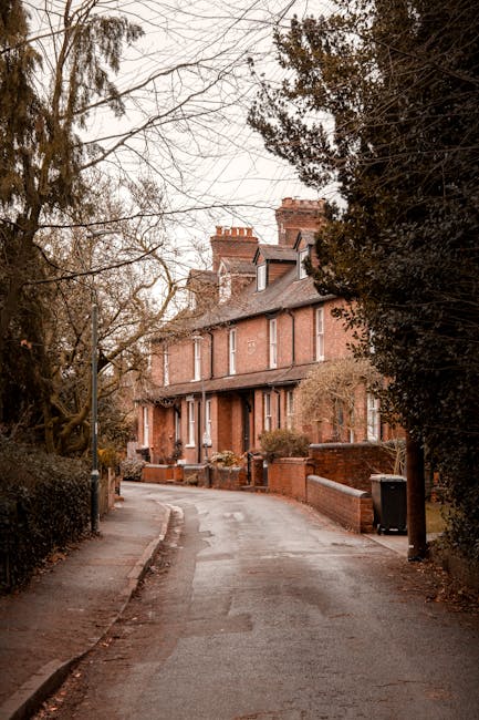 A narrow residential street in Hampstead lined with brick terraced houses featuring pitched roofs and chimneys, surrounded by leafless trees and overcast sky, with a pavement on one side and a small driveway leading to the houses. In the foreground, a black waste bin is positioned near a brick wall, and the street surface appears damp. This scene illustrates the typical setting for home relocation or furniture transport services, with Hampstead Movers potentially assisting with packing, loading, and moving household belongings through such narrow streets. The image captures the quiet, historic character of the area, emphasizing the challenges of house removals on tight, rural-style streets typical for London’s Hampstead neighborhood.