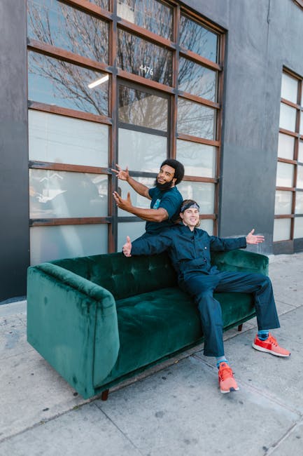 Two men are positioned outside a modern building with large glass windows framed by wooden panels. One man, standing behind a lush, deep green velvet sofa placed on the sidewalk, is smiling and gesturing towards the building, indicating a friendly interaction. The other man sits on the sofa with his arms extended outward, also smiling, and appears to be in a cheerful mood. The scene suggests a home relocation or furniture transport scenario, with the sofa possibly being prepared for moving or delivery. The sidewalk and exterior environment are well-lit, with natural daylight highlighting the scene. Hampstead Movers, a professional removals company, may be involved in this furniture transfer, highlighting their expertise in loading, packing, and transporting household items during house relocation tasks, especially on narrow streets like those on Rosslyn Hill. The image captures a moment of coordination or celebration during the furniture loading process related to house moving services.