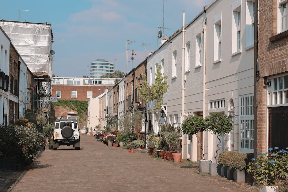 A narrow residential street in Hampstead lined with brick terraced houses featuring pitched roofs and chimneys, surrounded by leafless trees and overcast sky, with a pavement on one side and a small driveway leading to the houses. In the foreground, a black waste bin is positioned near a brick wall, and the street surface appears damp. This scene illustrates the typical setting for home relocation or furniture transport services, with Hampstead Movers potentially assisting with packing, loading, and moving household belongings through such narrow streets. The image captures the quiet, historic character of the area, emphasizing the challenges of house removals on tight, rural-style streets typical for London’s Hampstead neighborhood.