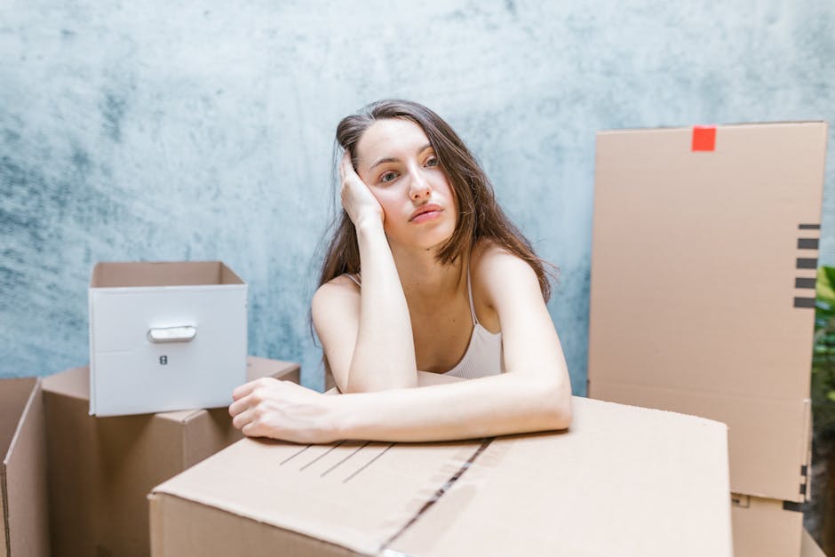A young woman with long brown hair and a neutral expression, resting her head on her left hand, sitting behind a stack of cardboard boxes during a home relocation or packing process. The scene is set indoors against a light blue textured wall, with various open and closed cardboard boxes around her, some sealed with red tape and others open revealing packing materials. She is wearing a sleeveless top, and her right arm rests on a large cardboard box in front of her. In the background, there is a white box with a handle and other storage containers. The environment suggests a packing or moving situation, which relates to furniture transport and home removals, noting Hampstead Movers’ services for professional removals and packing support.