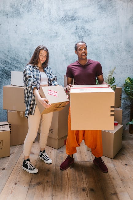A young woman with long brown hair and a neutral expression, resting her head on her left hand, sitting behind a stack of cardboard boxes during a home relocation or packing process. The scene is set indoors against a light blue textured wall, with various open and closed cardboard boxes around her, some sealed with red tape and others open revealing packing materials. She is wearing a sleeveless top, and her right arm rests on a large cardboard box in front of her. In the background, there is a white box with a handle and other storage containers. The environment suggests a packing or moving situation, which relates to furniture transport and home removals, noting Hampstead Movers’ services for professional removals and packing support.